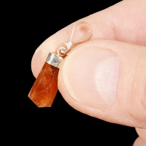 May include: A faceted, orange-brown gemstone pendant with a silver-colored bail. The pendant is held against a black background. The gemstone has a rectangular shape with angled sides and a polished finish. Jewelry.