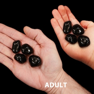 May include: A comparison of the size of black obsidian tumbled stones in the hands of an adult and a 7-year-old child.  The text "7 YR OLD" and "ADULT" is visible in the image.