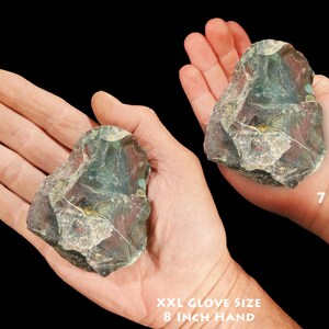 May include: Two hands holding a large, green, red, and white, irregularly shaped rock. The rock is about the size of a person's hand. The hand on the left is an adult hand, and the hand on the right is a child's hand. The text "XXL Glove Size 8 inch Hand" is below the adult hand, and the text "7 Year Old" is below the child's hand.