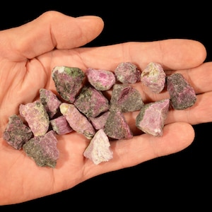 May include: A handful of rough ruby crystals, displaying a mix of pink, red, and green hues. The gemstones are held in an open hand against a black background, showcasing their natural, unpolished texture. These raw rubies are ideal for jewelry making or collecting.