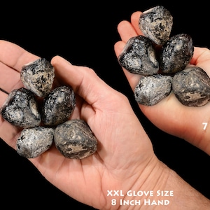 May include: A comparison of the size of black obsidian stones in the hands of a 7-year-old and an adult with an 8-inch hand. The stones are smooth and have a dark, glassy appearance.  "7 YEAR OLD" and "XXL GLOVE SIZE 8 INCH HAND" are written in white text on the image.