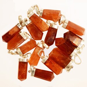 May include: A collection of orange-brown gemstone pendants with silver-colored metal caps and loops. The pendants are various sizes and shapes, with a faceted or polished appearance. The background is a plain white surface.