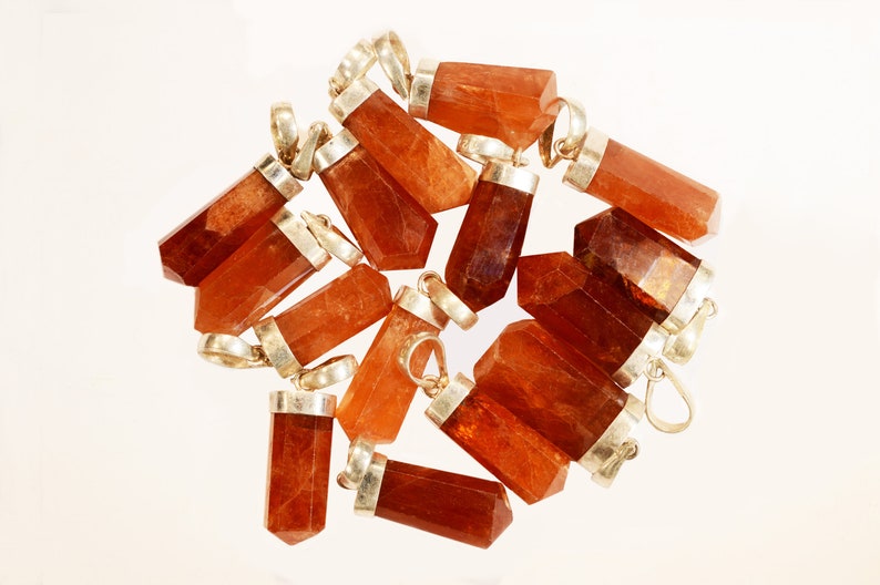 May include: A collection of orange-brown gemstone pendants with silver-colored metal caps and loops. The pendants are various sizes and shapes, with a faceted or polished appearance. The background is a plain white surface.