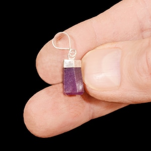 May include: A silver pendant with a rectangular purple gemstone. The pendant is held between two fingers.