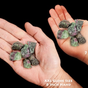 May include: A comparison of the size of green and pink rough ruby zoisite crystals in the hands of a 7-year-old and an adult with an 8-inch hand.  The crystals are about the size of a dime.