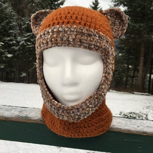 May include: A crocheted hat with brown and beige yarn, featuring two floppy ears. The hat is designed to cover the head and neck, with a fold-over flap at the neck.