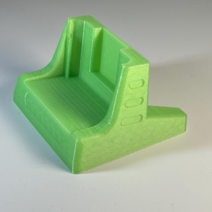 May include: A lime green 3D printed plastic bracket featuring a flat base, a vertical back support, and side reinforcements with three small oval indentations on each side. The object has a smooth finish with visible horizontal layer lines.