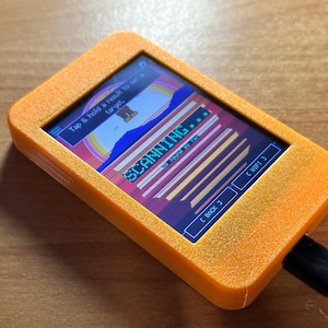 May include: A rectangular orange handheld electronic device with a digital screen displaying a colorful graphic interface. The screen shows the text SCANNING, Tap & hold a result to set a target, 0 found so far, C BACK, and C WIFI 3. A black cable is plugged into the side.