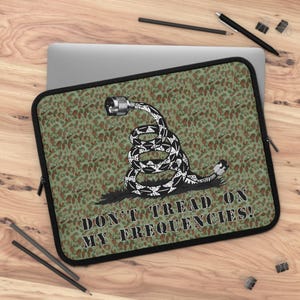 Puede incluir: Una funda para portátil de camuflaje verde y marrón con una serpiente negra enroscada alrededor de un cable y el texto "Don't Tread On My Frequencies!"