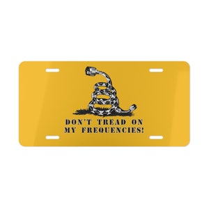 Puede incluir: Una placa de matrícula amarilla con una serpiente enroscada en blanco y negro con un conector de cable en la cabeza. El texto "DON'T TREAD ON MY FREQUENCIES!" está impreso debajo de la serpiente.