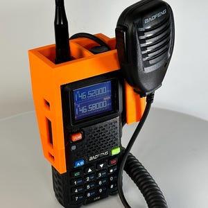 Può includere: Un supporto radio arancione stampato in 3D con una radio e un microfono Baofeng neri. La radio visualizza le informazioni sulla frequenza su uno schermo blu. Il supporto ha uno slot per l'antenna e una clip per il microfono.