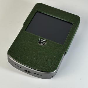 May include: A dark green electronic device with a black screen. The device has a textured surface and a silver-colored frame. A small camera lens is visible near the top of the screen.