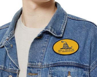 Könnte beinhalten: Eine blaue Jeansjacke mit einem gelben ovalen Aufnäher, auf dem "Don't Tread On My Frequencies!" steht und ein schwarz-weißes Schlangenmotiv mit einem eingerollten Schwanz zu sehen ist.