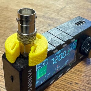 May include: A black radio receiver with a digital display showing frequency information. A silver antenna connector is attached, held in place by a bright yellow plastic holder. The device is labeled "S Mini V4".