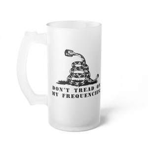 Puede incluir: Una jarra de cerveza de vidrio blanco esmerilado con una imagen en blanco y negro de una serpiente con un cable en espiral para la cola. La serpiente está sobre un fondo verde y el texto "Don't Tread On My Frequencies" está debajo de la serpiente.