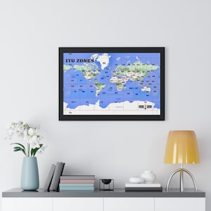 May include: A framed world map with a blue background and black grid lines. The map is labelled "ITU ZONES" and shows the different zones of the International Telecommunication Union. The map is hanging on a white wall above a shelf with books and a vase of white flowers.