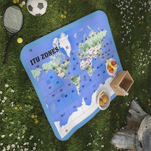 May include: A blue and white picnic blanket featuring a printed world map. The map is divided into zones labelled with numbers. The blanket is laid out on a grassy surface with a football, a small wooden box, and a plate of fruit.