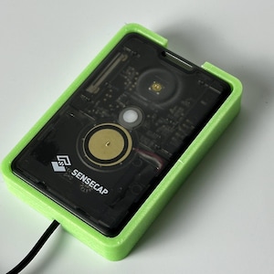 Może przedstawiać: Czarne urządzenie elektroniczne z widocznymi elementami wewnętrznymi jest zamknięte w jasnozielonej prostokątnej ramie. Urządzenie ma okrągły, złoty element, a widoczny jest tekst "SENSECAP". Czarny kabel wychodzi z urządzenia.