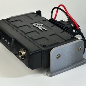 May include: A black electronic device with the "OVGC" logo, a gray mounting bracket, and attached red and black wires. The device has a textured surface and multiple ports. The bracket is secured with gold screws.
