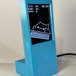 May include: A light blue, 3D-printed desktop monitor stand featuring a small vertical screen. The display shows system statistics including CPU: 0%, RAM: 0%, and WPM: 0, alongside a pixel art graphic of a creature typing on a keyboard.