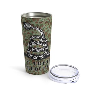 Puede incluir: Un vaso de viaje de acero inoxidable con un patrón de camuflaje verde, marrón y negro. El vaso presenta un gráfico de una serpiente enroscada con un conector al final de su cola. El texto "Don't Tread On Me" está impreso en el vaso.