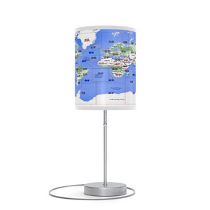May include: A silver table lamp with a white cylindrical shade featuring a world map with blue oceans and green continents. The map is divided into squares with numbers indicating latitude and longitude. The lamp has a silver base and a silver ring around the base.