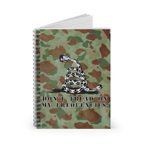 Puede incluir: Una libreta espiral de camuflaje verde con una ilustración en blanco y negro de una serpiente enroscada alrededor de un conector de cable. El texto "Don't Tread On My Frequencies!" está impreso debajo de la imagen.