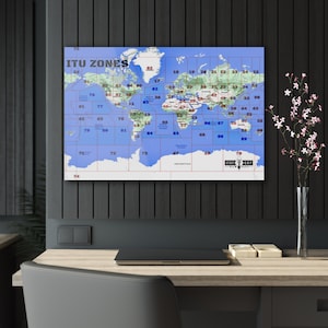 May include: A world map with blue and white colours showing the ITU zones. The map is divided into 79 zones, each with a number. The map is framed and hanging on a black wall.