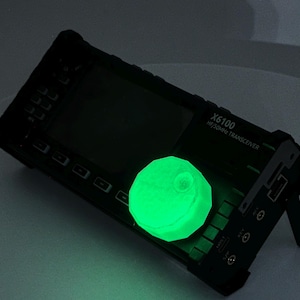 May include: A black X6100 HF/50MHz transceiver with a glowing green sphere on top. The device has a screen and several buttons. The sphere is emitting a bright green light, contrasting with the dark body of the transceiver.