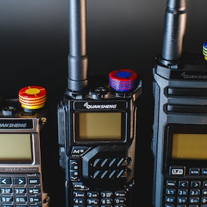 May include: Three black and silver QuanSheng walkie-talkies with antennas and colorful top knobs. The devices have keypads, screens, and the QuanSheng logo. The background is dark.