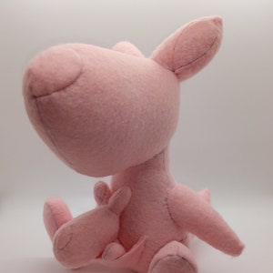 May include: A pink plush kangaroo toy with a joey in its pouch. The kangaroo is sitting on a white surface.
