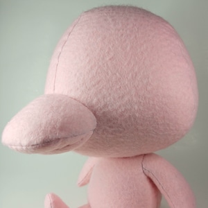 Puede incluir: Un juguete de peluche rosa con forma de pájaro con una cabeza grande y un pico pequeño. El juguete está sentado sobre una superficie blanca.