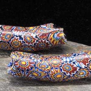 Puede incluir: Dos cuentas cilíndricas de vidrio con un complejo patrón millefiori de motivos repetidos en forma de estrella en tonos amarillo, azul, blanco y marrón rojizo. Las cuentas tienen una forma tubular ligeramente curva con un acabado suave.
