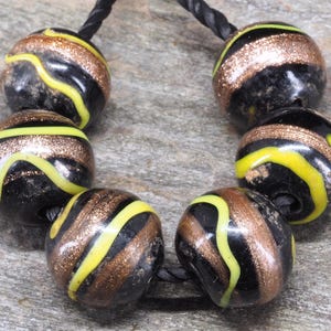 Puede incluir: Seis cuentas de vidrio esféricas ensartadas en un cordón negro. Cada cuenta es negra con una banda horizontal de purpurina brillante de color cobre y una fina línea amarilla ondulada que envuelve la superficie en un patrón repetitivo.
