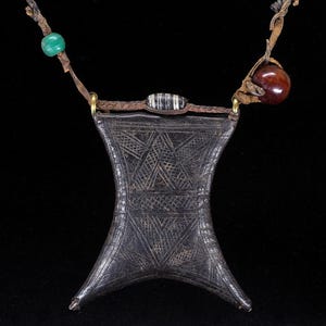 May include: A dark brown leather pouch with an hourglass shape and intricate geometric etched patterns. The pouch hangs from a braided leather cord adorned with a green bead, a dark red bead, and a small black and white striped cylindrical bead.