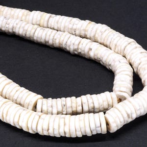 May include: Close-up of a necklace made of white, flat, circular beads. The beads are strung together, forming a flexible strand. The beads have a slightly textured surface and a natural, off-white color. The necklace is displayed against a dark background.