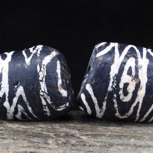 Puede incluir: Dos cuentas cilíndricas oscuras con una superficie negra mate desgastada, decoradas con intrincados patrones lineales blancos pintados a mano. Las cuentas presentan un acabado rugoso y texturizado con signos visibles de desgaste.
