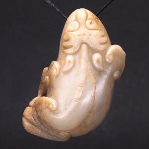 May include: Cream-colored carved jade pendant depicting a stylized animal with intricate details. The pendant is suspended from a black cord, showcasing the detailed craftsmanship and the smooth, polished surface of the jade.