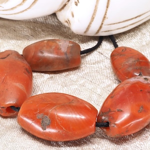 Peut inclure: Un collier composé de grandes perles ovales rouge-orangé enfilées sur un cordon noir. Les perles ont une finition polie et brillante avec des variations naturelles de couleur et de texture. Le collier est présenté sur un fond clair texturé.