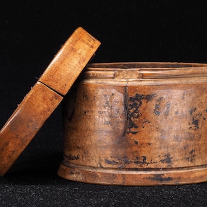 May include: A cylindrical wooden box with a separate lid leaning against its side. The wood features a weathered, light brown finish with dark, distressed patches and a visible vertical crack running down the front of the container body.