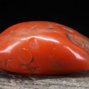 Peut inclure: Une pierre polie de forme ovale, d'une couleur rouge-orange vibrante. La pierre présente des imperfections naturelles et une finition brillante. Elle repose sur une surface grise texturée, sur fond sombre.
