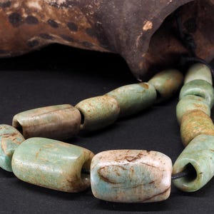Puede incluir: Un collar de cuentas antiguas en forma de barril, en tonos verdes y beige. Las cuentas están ensartadas en un cordón negro, con un objeto oscuro y texturizado en el fondo. Las cuentas tienen un aspecto envejecido.