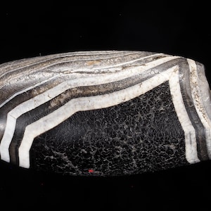 Puede incluir: Una cuenta cilíndrica con un centro negro prominente y una superficie texturizada y agrietada. La cuenta está decorada con bandas alternas blancas y gris oscuro con patrón de chevrón que envuelven ambos extremos, en un cordón fino.