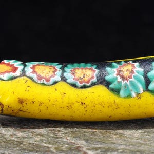 Puede incluir: Una cuenta de vidrio amarilla con un patrón floral. La cuenta tiene una forma rectangular curva con una base amarilla y una sección superior con un patrón floral repetido en rojo, naranja, verde y blanco.
