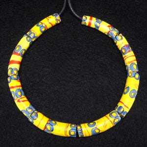 Puede incluir: Un collar con cuentas cilíndricas amarillas ensartadas en un cordón negro. Cada cuenta muestra un patrón repetitivo de motivos circulares azules y finas líneas horizontales rojas, creando un diseño vibrante en toda la pieza.