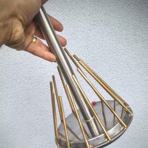 Könnte beinhalten: Ein Küchenutensil mit einem zentralen Griff aus Edelstahl, der an einer kreisförmigen Basis befestigt ist. Eine Reihe dünner, vertikaler Stäbe in Messingoptik ist um den Rand der Basis angeordnet und erstreckt sich nach oben zum Griff. Ein kleiner roter Knopf ist sichtbar.