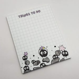 May include: A rectangular notepad with a white grid pattern and the text THINGS TO DO at the top. The bottom features illustrations of small, round, black fuzzy creatures with large eyes, sitting on gray rocks and holding colorful pastel stars.