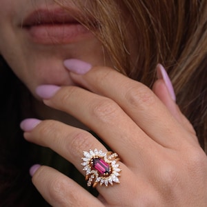 May include: A gold ring with a large, pink, rectangular gemstone surrounded by smaller, white gemstones. The ring is set in a delicate, floral design.