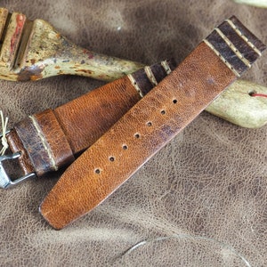 May include: A brown leather watch strap with white stitching and a silver buckle. The strap is lying on a brown leather surface with other leatherworking tools.