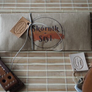 May include: A brown leather watch strap with stitching and two holes. The strap is packaged in a brown paper envelope with a string tied around it. The envelope has a brown leather tag with the text "Sk&oacute;rnik - Styl" on it.